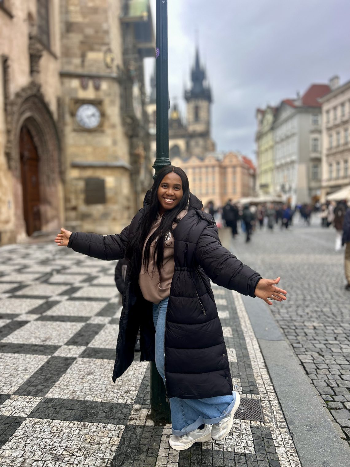 A picture of a lady with a blurred background standing in front of a lamp post with arms spread out, smiling.