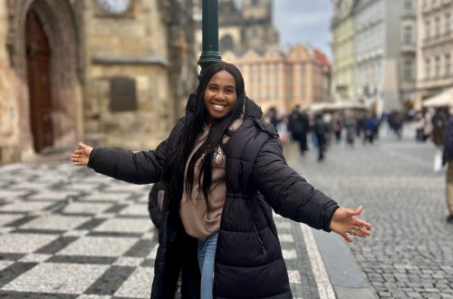 A picture of a lady with a blurred background standing in front of a lamp post with arms spread out, smiling.
